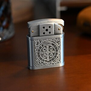 May include: A silver-colored metal lighter with an ornate design. The lighter features a detailed engraving with the text "PAX" and other symbols. The lighter is open, revealing the interior components. The background is a blurred wooden surface.