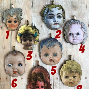 K&ouml;nnte beinhalten: Acht Vintage-Puppenkopf-Anh&auml;nger mit verschiedenen Frisuren und Gesichtsz&uuml;gen. Die Anh&auml;nger sind aus Holz gefertigt und haben einen Used-Look. Sie h&auml;ngen alle an einer Holzoberfl&auml;che.