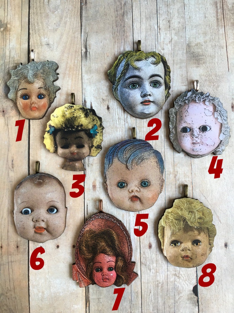 K&ouml;nnte beinhalten: Acht Vintage-Puppenkopf-Anh&auml;nger mit verschiedenen Frisuren und Gesichtsz&uuml;gen. Die Anh&auml;nger sind aus Holz gefertigt und haben einen Used-Look. Sie h&auml;ngen alle an einer Holzoberfl&auml;che.