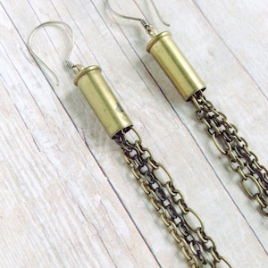 May include: A pair of brass-colored bullet casing earrings with silver-tone ear wires. Each earring features a cascade of layered, antique brass-tone chains. The earrings are displayed against a light wood-grain background.