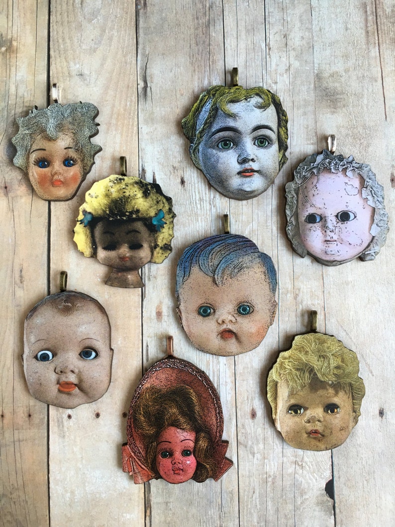 K&ouml;nnte beinhalten: Acht Vintage-Puppenkopf-Anh&auml;nger mit bemalten Gesichtern und Haaren. Die Anh&auml;nger sind aus Holz und haben einen Used-Look. Sie sind alle in verschiedenen Farben und haben verschiedene Frisuren.