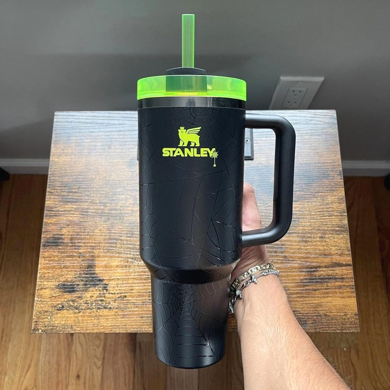 Op de afbeelding: Zwarte Stanley-beker met een neon groen deksel en rietje. De beker heeft een grote handgreep en een spinnenwebontwerp. Het Stanley-logo is in neon groen. De beker staat op een houten tafel.