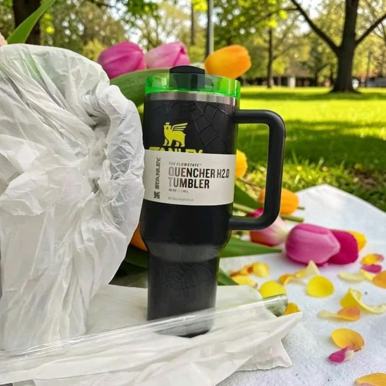 Op de afbeelding: Zwarte Stanley Quencher H2.0 tumbler met een groene deksel en handvat. De tumbler heeft een textuurontwerp en een wit label met de tekst "QUENCHER H2.0 TUMBLER". De tumbler staat op een wit oppervlak met bloemen op de achtergrond.