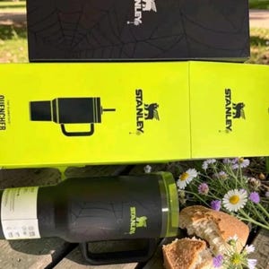 Op de afbeelding: Een zwarte Stanley Quencher tumbler met een handvat en een doorzichtig deksel, in de buurt van een doos met het Stanley-logo. De tumbler staat op een houten oppervlak met bloemen en brood.