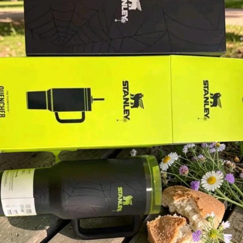Op de afbeelding: Een zwarte Stanley Quencher tumbler met een handvat en een doorzichtig deksel, in de buurt van een doos met het Stanley-logo. De tumbler staat op een houten oppervlak met bloemen en brood.