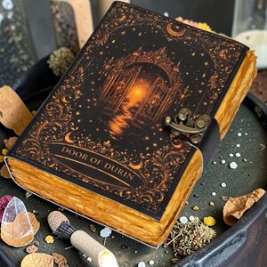 以下が含まれることがあります： 「DOOR OF DURIN」のテキストと装飾的なデザインが施された黒い表紙の装飾ジャーナル。ジャーナルは、古くなった黄ばんだページと金属製の留め金が付いています。表紙のデザインには、門、星、三日月が含まれています。