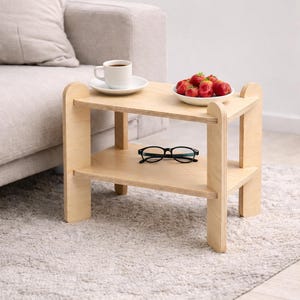 May include: A small, light-colored wooden side table with two shelves. A white plate of strawberries, a cup of coffee, and a pair of black glasses rest on the table. The table is next to a gray sofa.