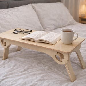 Könnte beinhalten: Ein hellfarbiger Betttablett aus Holz mit Klappbeinen, auf dem ein Buch, eine Brille und eine weiße Tasse liegen. Das Tablett hat dekorative Ausschnitte an den Seiten und steht auf einem Bett mit weißer Bettwäsche.