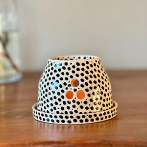 May include: A ceramic oil burner with a white base and black polka dot pattern. The piece features a wide, tapered body with three circular cutouts arranged in a triangle on the front, revealing an orange interior, and sits on a matching saucer.