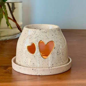 May include: A cream-colored ceramic wax warmer with a speckled finish and a matching circular base. The dome-shaped body features two heart-shaped cutouts, one smaller than the other, revealing a warm orange glow from within the hollow interior.