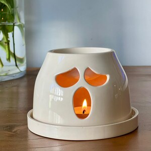 May include: A cream-colored ceramic wax warmer shaped like a ghost with cutouts for eyes and a mouth. The hollow interior contains a lit tea light candle, casting a warm orange glow through the openings. The piece sits on a circular base.