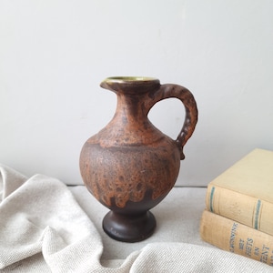 Op de afbeelding: Een keramische kan met een bolvormig lichaam en een voetstuk. Het oppervlak vertoont een gevlekt bruin en beige glazuur met een druppelpatroon. De kan heeft een enkel gebogen handvat aan de zijkant en een brede, licht uitlopende rand.