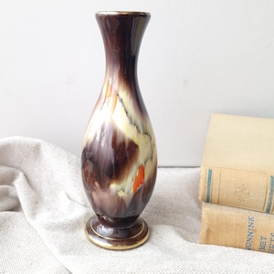 Op de afbeelding: Een keramische vaas met een glanzende afwerking en een gemarmerd patroon van donkerbruine, crèmekleurige en feloranje strepen. De vaas heeft een slanke hals, een bolvormig lichaam en een ronde voet met een dunne goudkleurige rand aan de boven- en onderkant.