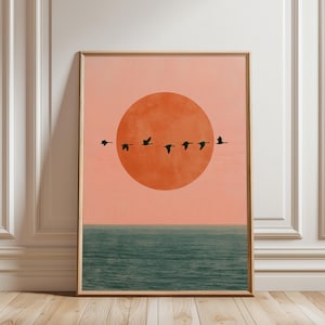 Könnte beinhalten: Ein rechteckiger Wandkunstdruck mit einer großen orangefarbenen Sonne vor einem sanften pfirsichfarbenen Himmel. Eine Gruppe von sechs schwarzen Vögeln in Silhouette fliegt horizontal vor der Sonne. Unter dem Himmel ein dunkelblaugrüner Ozean.