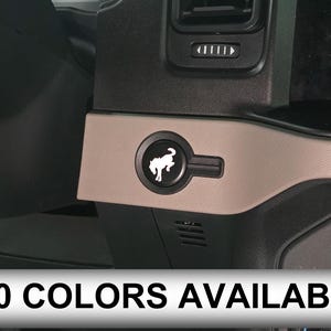 Op de afbeelding: Een beige dashboardpaneel met een ronde uitsparing voor een startknop, voorzien van een wit logo van een steigerend paard. Het paneel is gemonteerd op een zwart interieurpaneel van een voertuig. De tekst 110 COLORS AVAILABLE is zichtbaar.