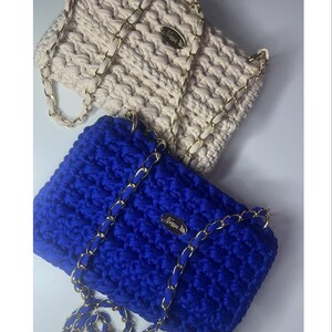 Peut inclure: Deux sacs en crochet faits à la main. L'un est de couleur crème, l'autre bleu roi. Les deux ont des chaînes dorées et une petite étiquette dorée avec l'inscription "Nessa". Les sacs sont rectangulaires avec un motif de point texturé.