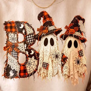 May include: A beige crewneck sweatshirt featuring a graphic print of the word BOO. The letter B is composed of various orange, black, and white plaid and lace patches. Two ghosts wearing orange and black plaid witch hats with small pumpkins.