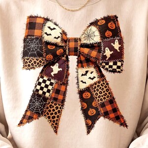 May include: A beige crewneck sweatshirt featuring a large, central graphic of a bow made from a patchwork of Halloween-themed fabrics. The patterns include orange and black buffalo plaid, spider webs, pumpkins, ghosts, bats, and leopard print.