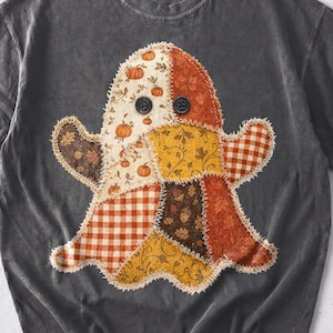 May include: A charcoal gray short-sleeve t-shirt featuring a central ghost-shaped graphic made of various patchwork fabric patterns in orange, brown, and cream colors, including pumpkins, leaves, and plaid, with two black buttons as eyes.