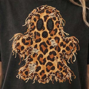 Może przedstawiać: Czarny t-shirt z centralną grafiką w kształcie ducha z brązowo-czarnym wzorem w panterkę. Wzór ducha ma postrzępione krawędzie, widoczne detale szwów, dwoje czarnych owalnych oczu oraz małe czarne okrągłe usta.