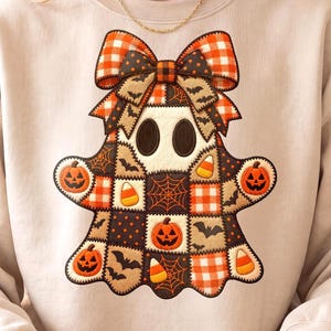 May include: A beige crewneck sweatshirt featuring a large, central graphic of a ghost made of a patchwork quilt design. The ghost is decorated with orange and black patterns, including pumpkins, bats, spiderwebs, candy corn, and a large bow.