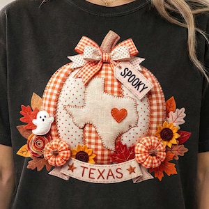 May include: A black t-shirt featuring a graphic of a pumpkin with orange and white gingham patterns. The pumpkin has a white Texas-shaped patch with a small orange heart, a bow, and a tag reading SPOOKY. A banner at the bottom reads TEXAS.
