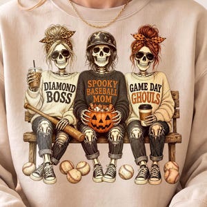 May include: A beige crewneck sweatshirt featuring a graphic of three skeletons sitting on a bench. The skeletons wear casual clothing and accessories, with text reading Diamond Boss, Spooky Baseball Mom, and Game Day Ghouls, plus a pumpkin bucket.