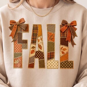 Könnte beinhalten: Ein beigefarbenes Sweatshirt mit Rundhalsausschnitt und dem Wort FALL in großen Blockbuchstaben. Jeder Buchstabe ist mit einem Patchwork aus herbstlichen Stoffmustern gefüllt, darunter Karos, Punkte und Blattmotive in Orange, Braun und Grün. Zwei braune Schleifen sind an den oberen Ecken der Buchstaben F und L befestigt.