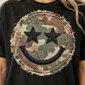 Może przedstawiać: Czarny t-shirt z krótkim rękawem z dużą okrągłą naszywką na piersi. Naszywka ma wzór moro z postrzępionymi brzegami, dwa czarne oczy w kształcie gwiazdek oraz czarny zakrzywiony uśmiech, tworząc wzór uśmiechniętej buźki.