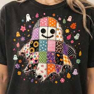 May include: A black t-shirt featuring a central graphic of a ghost made of various colorful patchwork squares, including floral, leopard print, and gingham patterns. The ghost has two black button eyes and a stitched mouth, surrounded by small ghosts, flowers, and leaves.