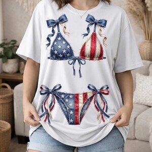 Può includere: Una t-shirt bianca a maniche corte con una stampa grafica di un bikini a tema patriottico. Il top e lo slip del bikini presentano una texture simile al denim, stelle bianche su sfondo blu e strisce verticali rosse e bianche.