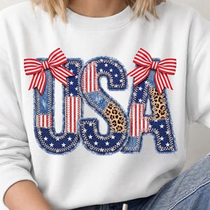 May include: White sweatshirt with the word "USA" in a patchwork design. The letters feature denim, stars, stripes, and leopard print, with red and white striped bows. The design is stitched with white thread.