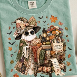 May include: A sage green t-shirt featuring a graphic of a ghost wearing a patterned witch hat and round glasses, pushing a shopping cart filled with pumpkins, lanterns, flowers, and a tote bag labeled Fall Finds Market with a receipt attached.