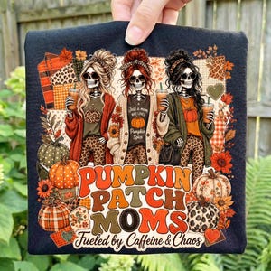 May include: A black t-shirt featuring a graphic of three skeletons with long hair and sunglasses, each holding a coffee cup. The text reads PUMPKIN PATCH MOMS Fueled by Caffeine & Chaos, surrounded by pumpkins, autumn leaves, and leopard print patterns.