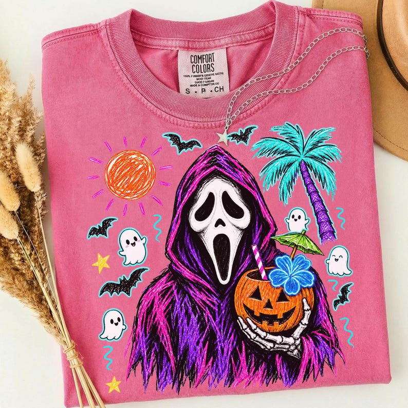 May include: A pink short-sleeve t-shirt featuring a graphic of a hooded figure in a purple robe holding a jack-o'-lantern with a straw and umbrella. The design includes a sun, palm tree, bats, and ghosts in a colorful, hand-drawn style.