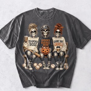 May include: A charcoal gray t-shirt featuring a graphic of three skeletons sitting on a bench. The skeletons wear baseball-themed clothing and accessories, with text that reads Diamond Boss, Spooky Baseball Mom, and Game Day Ghouls.