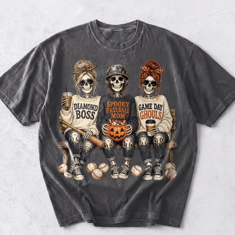 May include: A charcoal gray t-shirt featuring a graphic of three skeletons sitting on a bench. The skeletons wear baseball-themed clothing and accessories, with text that reads Diamond Boss, Spooky Baseball Mom, and Game Day Ghouls.