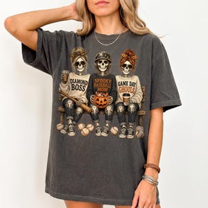 May include: A charcoal gray t-shirt featuring a graphic of three skeletons sitting on a bench. The skeletons wear sweaters with the text Diamond Boss, Spooky Baseball Mom, and Game Day Ghouls. They hold a baseball bat, a pumpkin, and a coffee cup.
