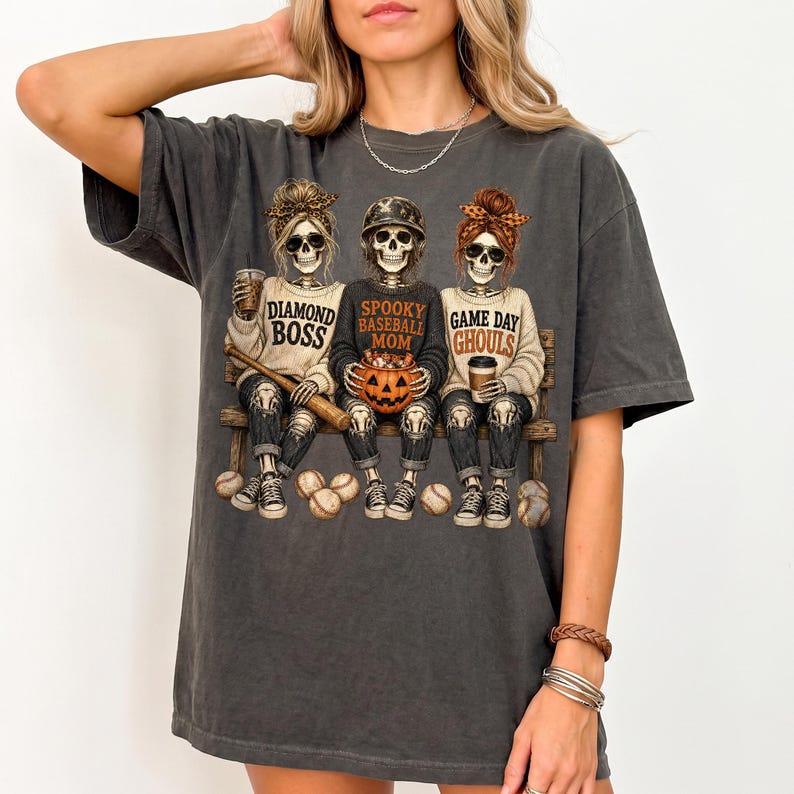 May include: A charcoal gray t-shirt featuring a graphic of three skeletons sitting on a bench. The skeletons wear sweaters with the text Diamond Boss, Spooky Baseball Mom, and Game Day Ghouls. They hold a baseball bat, a pumpkin, and a coffee cup.