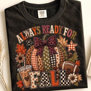 May include: A black t-shirt featuring a graphic print with the text ALWAYS READY FOR FALL. The design includes a patchwork pumpkin with a purple bow, a coffee cup, a football, autumn leaves, flowers, and stars in various patterns and colours.