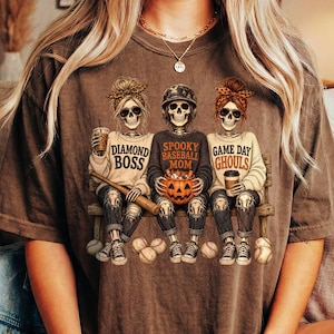 May include: A brown short-sleeve t-shirt featuring a graphic of three skeletons sitting on a bench. The skeletons wear sweaters with the text Diamond Boss, Spooky Baseball Mom, and Game Day Ghouls. They hold a baseball bat, a pumpkin, and a coffee cup.