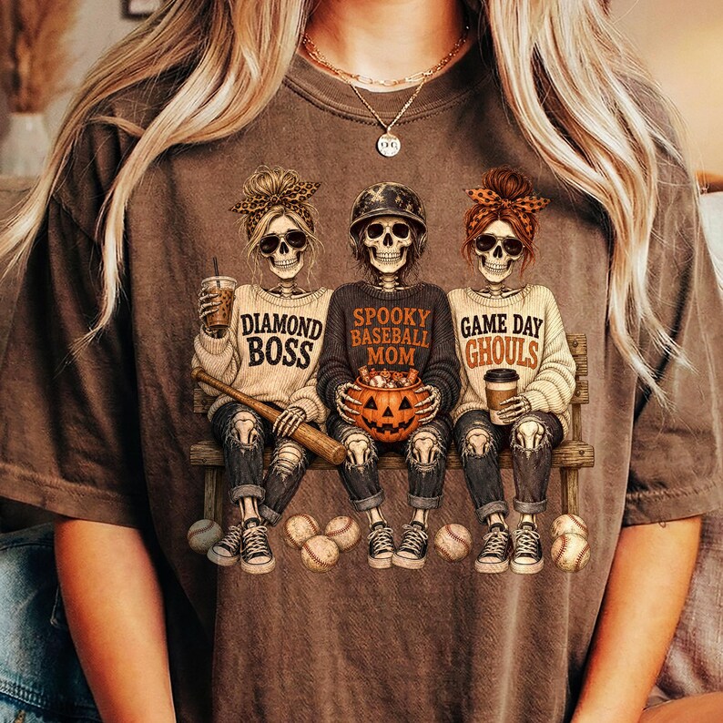 May include: A brown short-sleeve t-shirt featuring a graphic of three skeletons sitting on a bench. The skeletons wear sweaters with the text Diamond Boss, Spooky Baseball Mom, and Game Day Ghouls. They hold a baseball bat, a pumpkin, and a coffee cup.