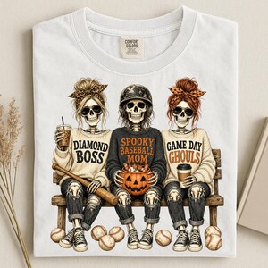May include: A white t-shirt featuring a graphic of three skeletons sitting on a bench. The left skeleton wears a leopard print headband and holds a drink. The center skeleton wears a baseball helmet and holds a pumpkin. The right skeleton wears an orange headband and holds a coffee cup. Text reads DIAMOND BOSS, SPOOKY BASEBALL MOM, and GAME DAY GHOULS.