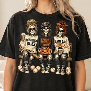 May include: A black t-shirt featuring a graphic of three skeletons sitting on a bench. The left skeleton wears a leopard print headband and holds a drink, with text reading DIAMOND BOSS. The center skeleton wears a baseball helmet, holds a pumpkin, and wears a shirt reading SPOOKY BASEBALL MOM. The right skeleton wears an orange headband and holds a drink, with text reading GAME DAY GHOULS.