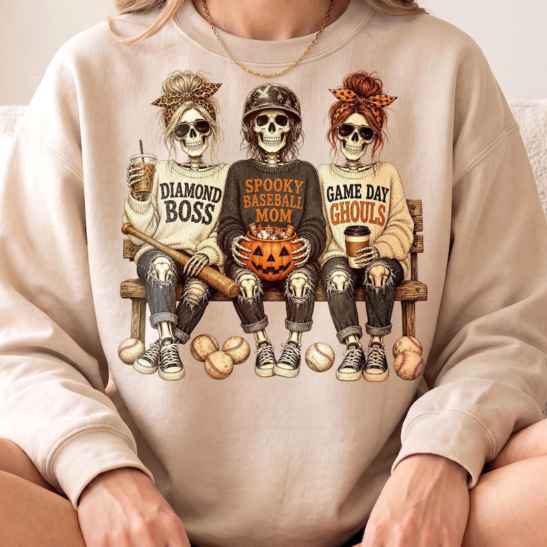 May include: A beige crewneck sweatshirt featuring a graphic of three skeletons sitting on a bench. The skeletons wear casual clothing and accessories, with text reading Diamond Boss, Spooky Baseball Mom, and Game Day Ghouls, plus a pumpkin bucket.