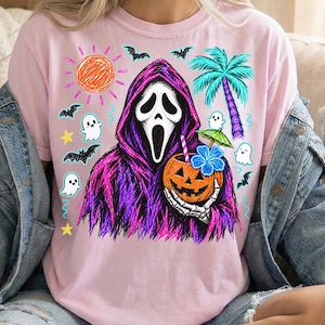 May include: A light pink t-shirt featuring a graphic of a hooded figure with a white mask holding a carved pumpkin with a straw and a blue flower. The design includes a bright orange sun, a teal palm tree, small white ghosts, bats, and yellow stars.