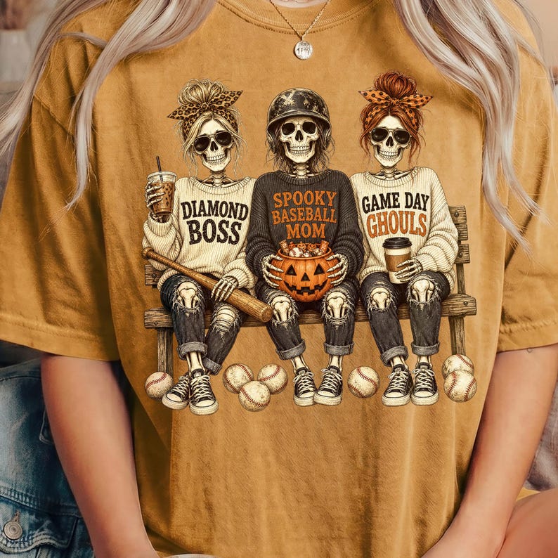 May include: A mustard yellow t-shirt featuring a graphic of three skeletons sitting on a bench. The left skeleton wears a leopard print bow and holds a drink, the center wears a baseball helmet and holds a pumpkin, and the right wears a bow and holds a drink. Text reads: DIAMOND BOSS, SPOOKY BASEBALL MOM, GAME DAY GHOULS.