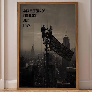 May include: A framed vertical wall art print featuring a dark, monochromatic cityscape with two figures perched atop a high antenna structure. The text reads 443 METERS OF COURAGE AND LOVE. The print is encased in a simple, light-toned wooden frame.