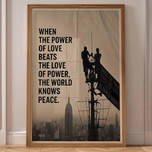 May include: A framed vertical poster featuring a silhouette of two figures atop a skyscraper antenna against a city skyline. The text reads: WHEN THE POWER OF LOVE BEATS THE LOVE OF POWER, THE WORLD KNOWS PEACE. The print has a light brown wooden frame.