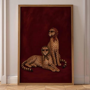 Op de afbeelding: Een ingelijste verticale kunstprint met twee cheeta's tegen een dieprode achtergrond. Beide cheeta's dragen hoofddoeken met patronen en een donkere zonnebril. De ene cheeta zit rechtop, terwijl de andere cheeta er voor op de grond ligt.